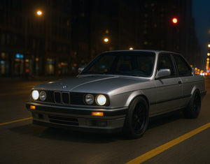 A gray BMW E30 driving through the city at night with AKG performance parts.